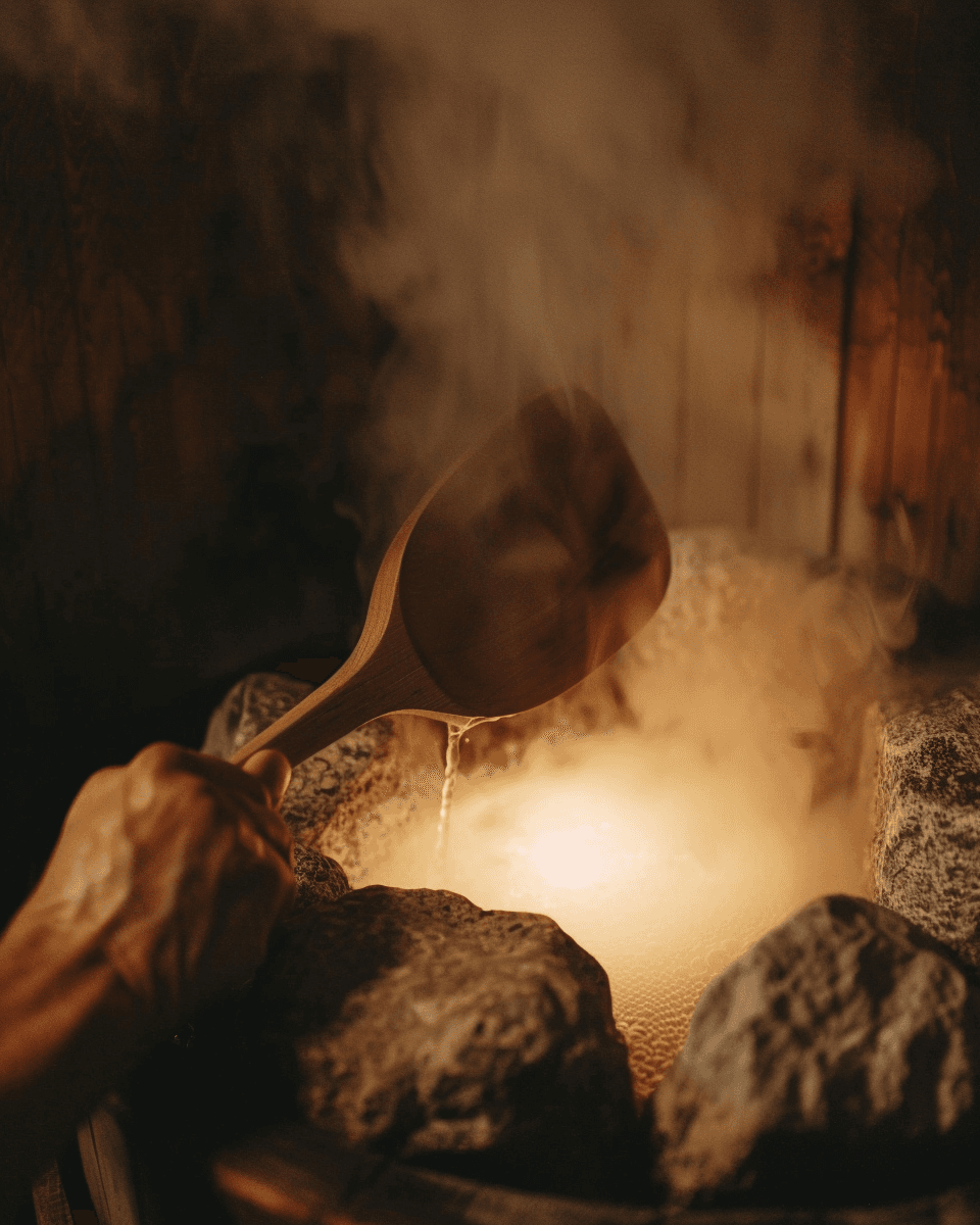 Hand holding a wooden ladle pouring water onto hot sauna stones creating löyly steam